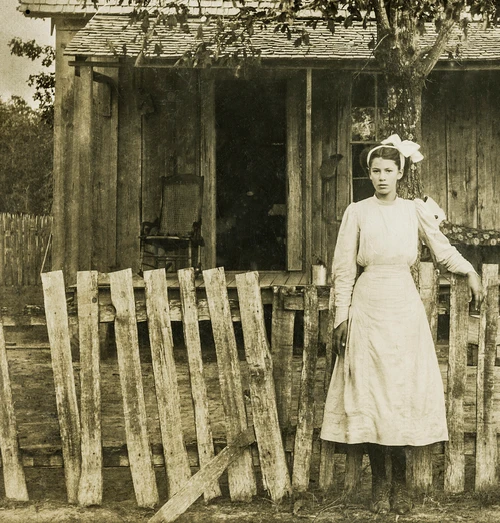 Antique photograph of a pioneer woman standing beside a weathered homestead